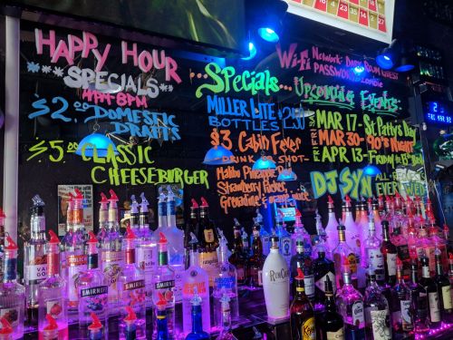 Colorful bar specials board featuring happy hour deals, drink prices, and upcoming events, with a backdrop of liquor bottles.
