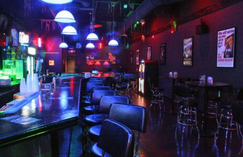 A dimly lit bar with colorful lights, empty stools, and a pool table, featuring posters on the walls.