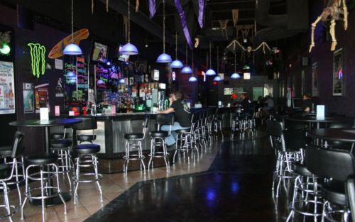 A dimly lit bar with blue pendant lights, high stools, and a long counter, featuring a vibrant drink selection.