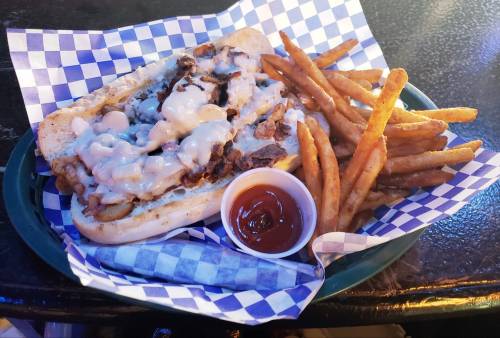 A loaded cheesesteak sandwich with fries and a side of dipping sauce on a checkered paper-lined plate.