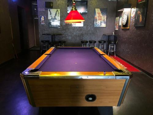 A pool table with a purple felt surface, under a red lamp, in a dimly lit room with bar stools and posters on the walls.