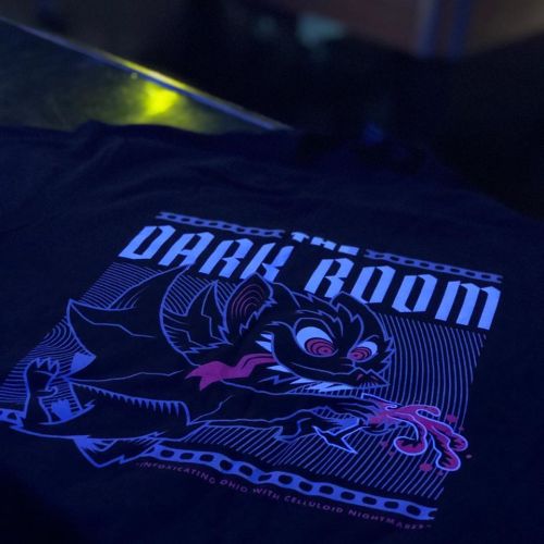 A black t-shirt featuring a graphic design of a creature with a sinister expression and the text "The Dark Room."