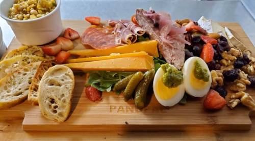 A wooden platter filled with cheeses, meats, pickles, boiled eggs, fruits, nuts, and a side of corn salad.