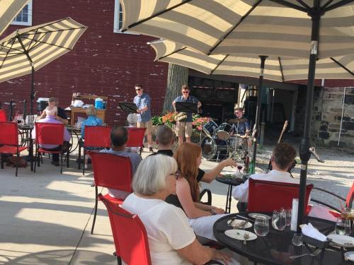 A live band performs outdoors under umbrellas, with an audience seated at tables enjoying food and drinks.