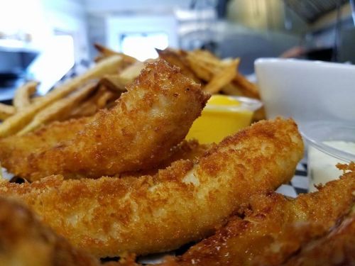 Close-up of crispy fried fish fillets with dipping sauces and golden fries in the background.