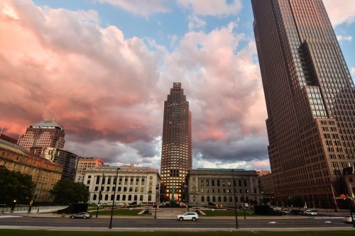 City skyline at sunset with dramatic clouds, featuring tall buildings and a historic structure in the foreground.