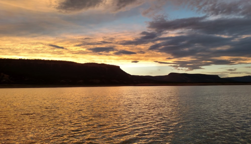 A serene sunset over a calm lake, with vibrant orange and purple hues reflecting on the water and silhouetted hills in the background.