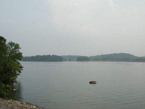 A calm lake surrounded by trees and hills, with a small floating platform in the water under a hazy sky.