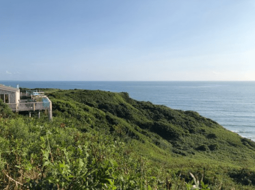 A coastal view featuring lush green hills and the ocean under a clear blue sky. A deck overlooks the scenery.