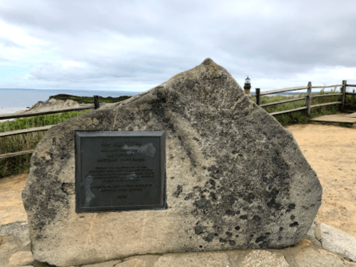 A large stone monument with a plaque, overlooking a coastal view and a lighthouse in the distance.