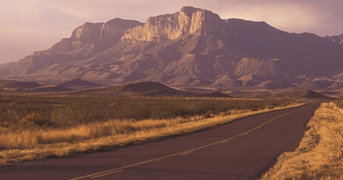 A winding road leads towards a majestic mountain range under a soft, cloudy sky.