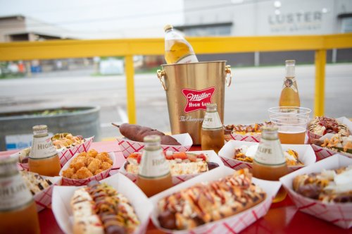 A table filled with various hot dogs, snacks, and beers in a bucket, set against an industrial background.
