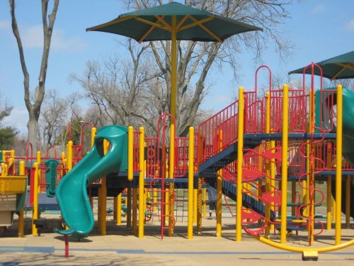 Colorful playground equipment with a green slide, climbing structures, and shaded areas under blue skies.