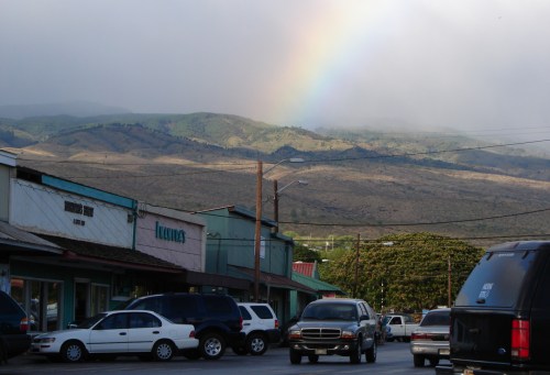 A rainbow arcs over green mountains, with a small town and parked cars in the foreground under a cloudy sky.