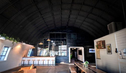 Interior of a modern, spacious café with a high arched ceiling, wooden accents, and a bar area with stools.