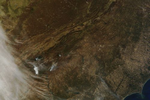Satellite image showing a vast landscape with varied terrain, including mountains, valleys, and some smoke plumes.