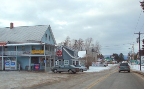 A snowy street scene featuring a general store and nearby buildings, with cars parked along the road.