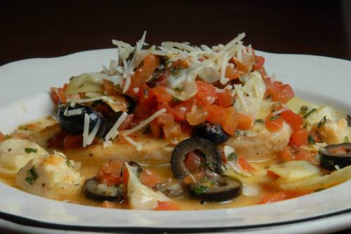 A plate of grilled fish topped with diced tomatoes, olives, and shredded cheese, garnished with herbs.