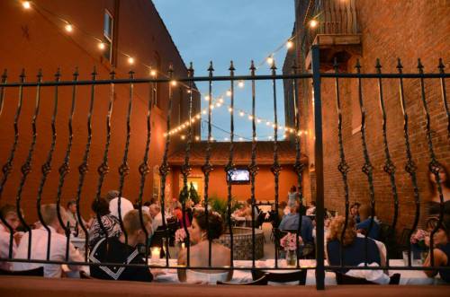 Outdoor dining area with string lights, featuring guests at tables and a cozy atmosphere in a brick-walled setting.