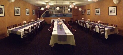A spacious dining room with long tables set for a gathering, decorated with flowers and a mural of a canal.