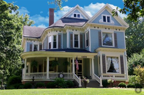 A large, elegant Victorian-style house with a wraparound porch, surrounded by greenery and under a blue sky.