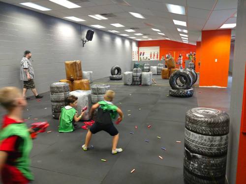 Children playing in a colorful indoor arena with tires and obstacles, engaged in a game with toy blasters.