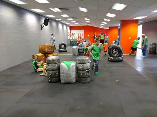 A vibrant indoor arena with players in green shirts engaging in a game, surrounded by stacked tires and obstacles.