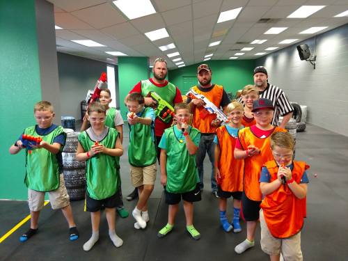 A group of kids and adults in colorful vests, holding toy blasters, posing in a game arena.