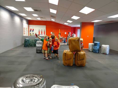 A group of children and an adult pose in a colorful play area with foam blocks and obstacles for games.