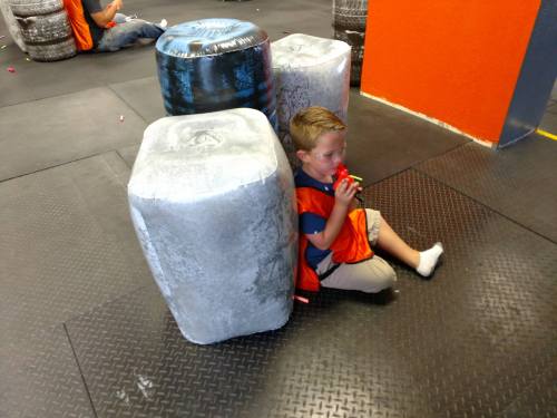 A young boy in an orange vest sits behind a foam barrier, holding a toy, in a play area with other kids in the background.