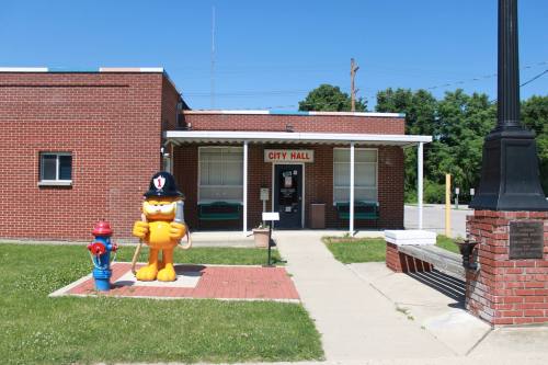 A brick city hall building with a cartoon firefighter statue and a fire hydrant in front on a sunny day.