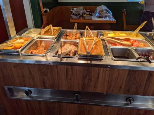 A buffet with various food trays, including meats, vegetables, and sides, displayed on a wooden counter.