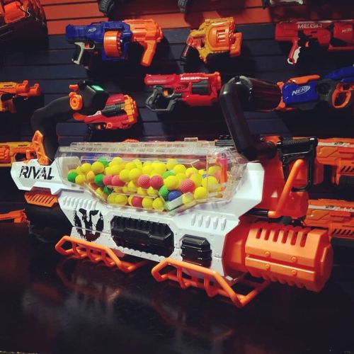 A colorful toy blaster filled with small foam balls, displayed against a backdrop of various other toy guns.