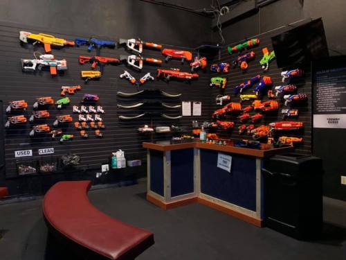 A wall display of various toy blasters and foam dart guns, with a counter and seating area in a play space.