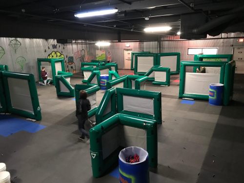 An indoor arena with inflatable barriers for a laser tag or paintball game, featuring blue barrels and a dark atmosphere.