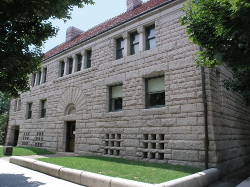 Historic stone building with a textured facade, arched entrance, and large windows, surrounded by greenery.