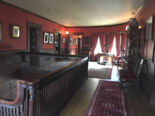 A warmly lit hallway with red walls, wooden railings, and vintage furniture, featuring framed pictures and a large window.
