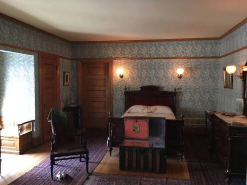 A vintage bedroom with patterned wallpaper, a wooden bed, a trunk, and antique furniture, softly lit by wall sconces.