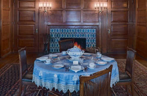 A beautifully set dining table with blue tablecloth, fine china, and a fireplace in a wooden-paneled room.
