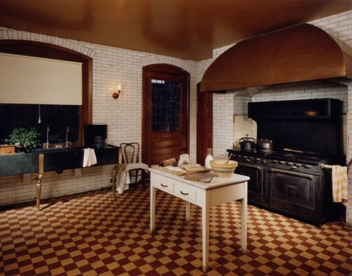 A vintage kitchen featuring a black stove, white table, checkered floor, and brick walls with a large hood above the stove.