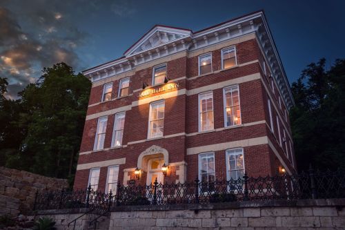 Historic brick building with large windows, illuminated at dusk, surrounded by trees and a decorative fence.