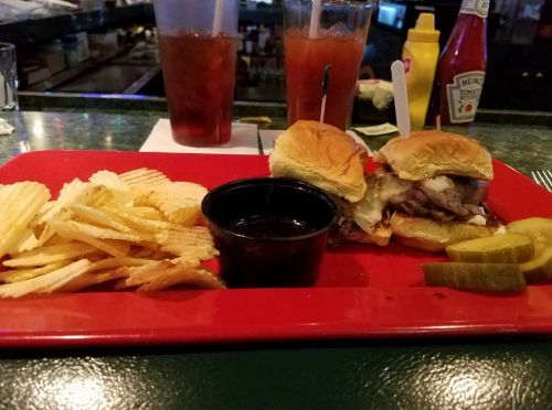 A red plate with two sliders, a small cup of sauce, potato chips, pickles, and drinks in the background.