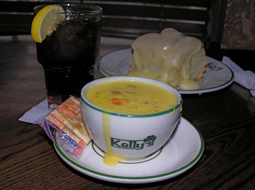 A bowl of creamy soup with vegetables, crackers, a glass of iced drink with lemon, and a dessert in the background.