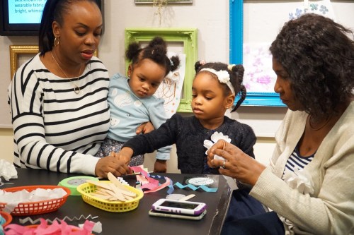 A woman and a girl craft together at a table, while another woman holds a toddler nearby. Colorful materials are scattered around.