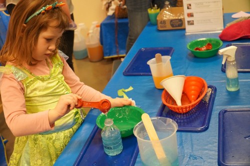 A young girl in a green dress measures liquid with a spoon at a colorful science activity table.