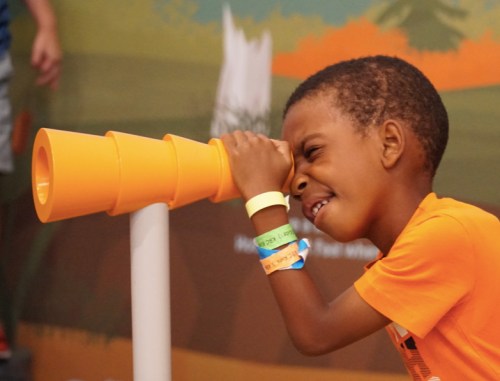 A young boy in an orange shirt looks through a bright orange telescope, smiling with excitement.
