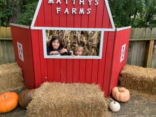 Two children peek out from a red barn structure surrounded by hay bales and pumpkins at a farm.