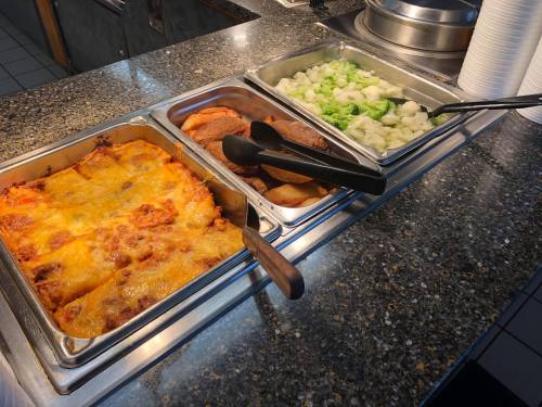 Three food trays on a buffet: one with cheesy lasagna, one with sliced meats, and one with steamed vegetables.
