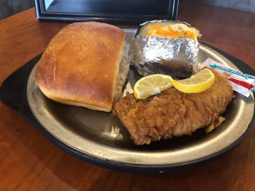 A plate with a piece of fried fish, a baked potato topped with cheese, a roll, and lemon slices.