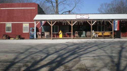 A rustic storefront with a red exterior, covered porch, and colorful chairs, set against a clear blue sky.